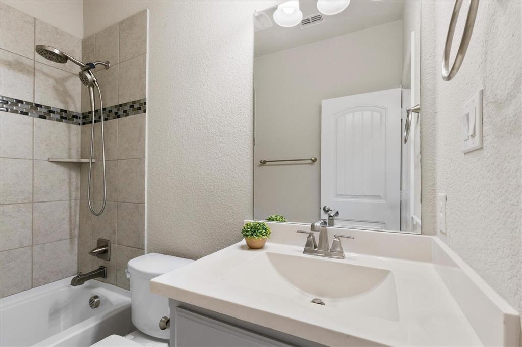 Vanity with a solid-surface countertop and integrated sink, complemented by a handheld shower fixture, tiled tub surround with decorative mosaic accent, and a full-width wall-mounted mirror