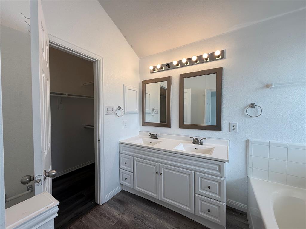 Dual-sink vanity with white cabinetry and solid-surface countertop, twin rectangular mirrors, multi-bulb vanity lighting, and wood-finish flooring