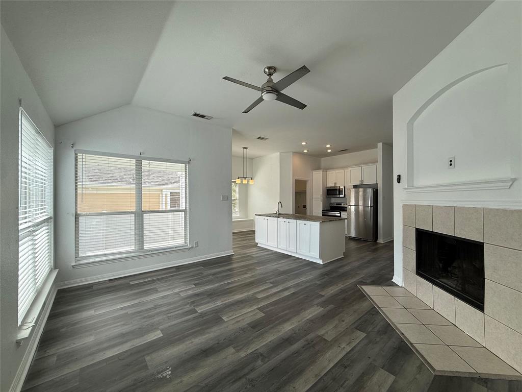Vaulted ceiling with an integrated ceiling fan, wood-finish flooring, tile-surround fireplace with a hearth, kitchen island with a neutral countertop, and white cabinetry