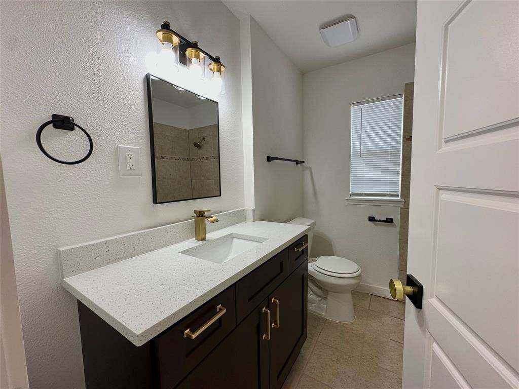 Bathroom featuring a vanity with quartz countertop, under-mount rectangular sink, brass-finish faucet, dark-finish cabinetry, and a rectangular mirror with integrated light fixture