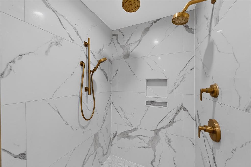 White and grey veined tile shower walls, dual shower heads, built-in recessed shelving, gold-finish fixtures, and a handheld shower