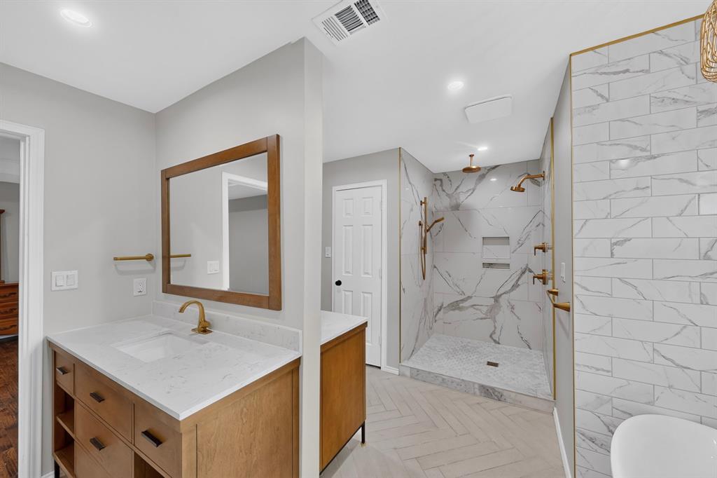 Dual shower system, wood-finish double sided vanity with countertop, framed mirror, herringbone pattern flooring, and white tiled shower walls
