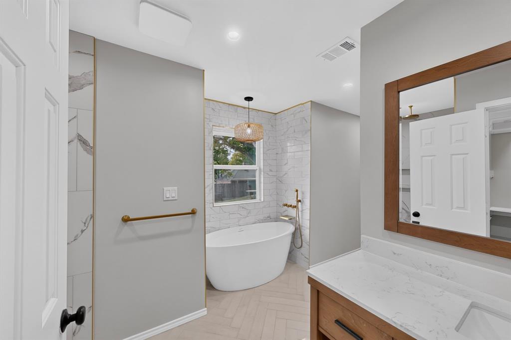 Freestanding soaking tub with gold-tone fixtures, white subway tile wall surround, and wood-finish vanity with a single basin and white countertop