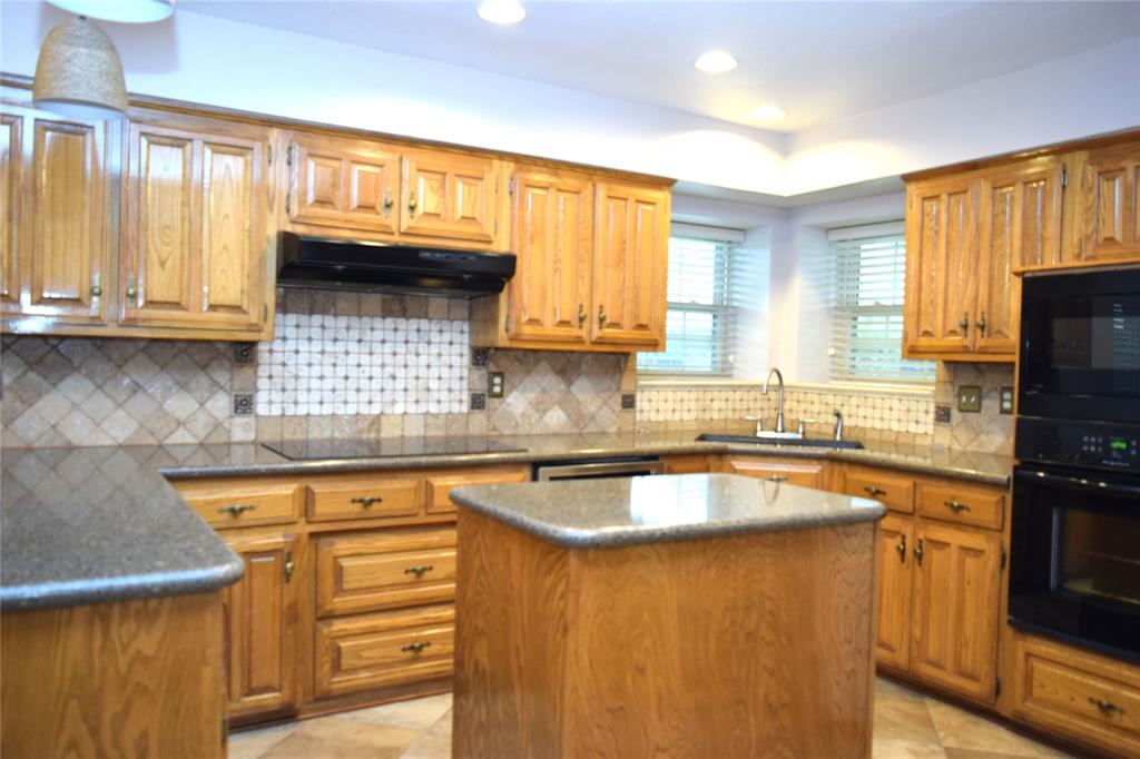 Kitchen featuring wood cabinetry, stone-finish countertops, a central island, tile backsplash with inset accent detailing, and built-in black wall ovens