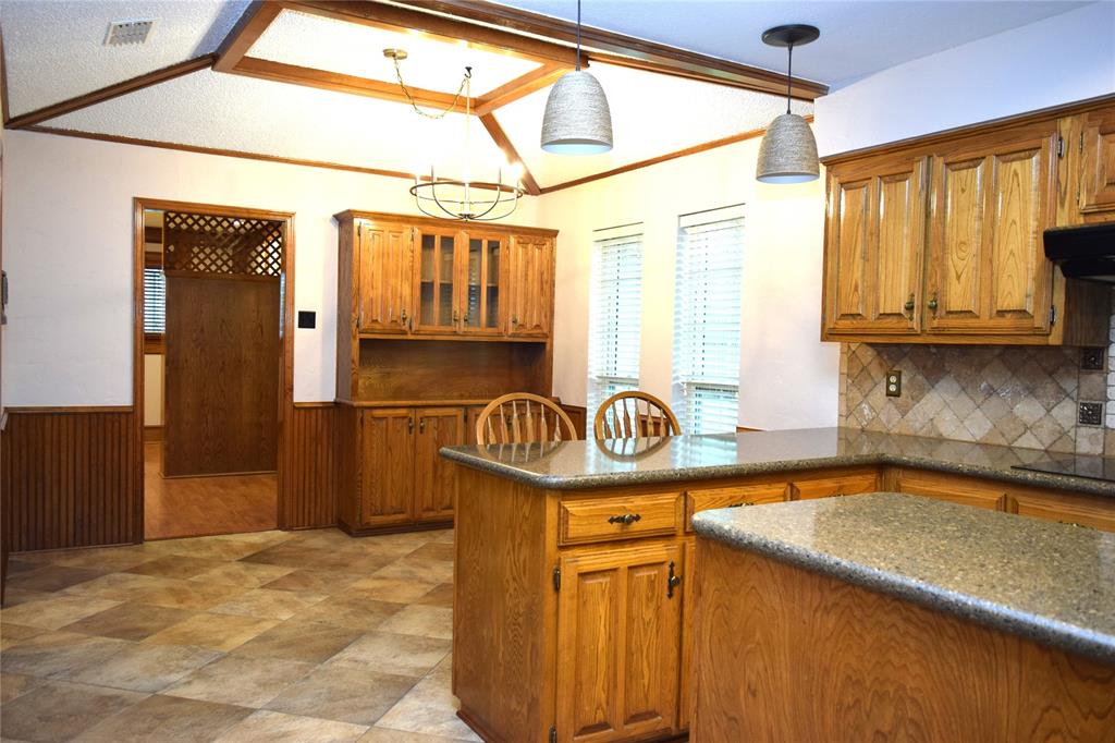 Kitchen featuring wood cabinetry, stone-finish countertops, tiled backsplash, vaulted ceiling with wood-beam detailing, and pendant lighting