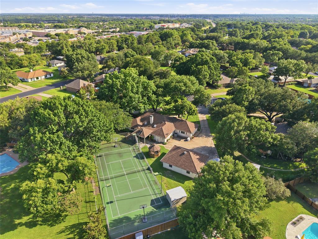 Aerial perspective of a sprawling property featuring a private tennis court, detached secondary structure, expansive mature tree canopy, and paved driveway