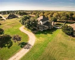 Aerial perspective of a multi-story residence featuring a winding driveway, expansive acreage, mature trees, and a covered porch