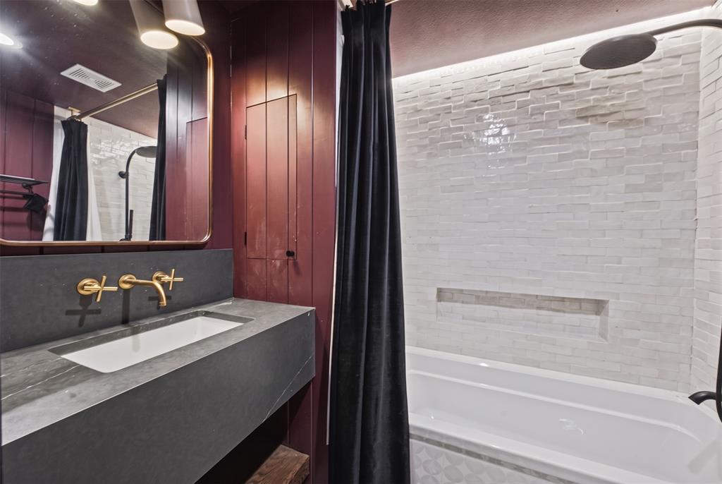 Floating vanity featuring a matte-finish dark countertop and integrated rectangular sink, accented by metallic wall-mounted widespread faucets, a textured white tile shower surround with a built-in recessed niche, and a metallic-framed vanity mirror