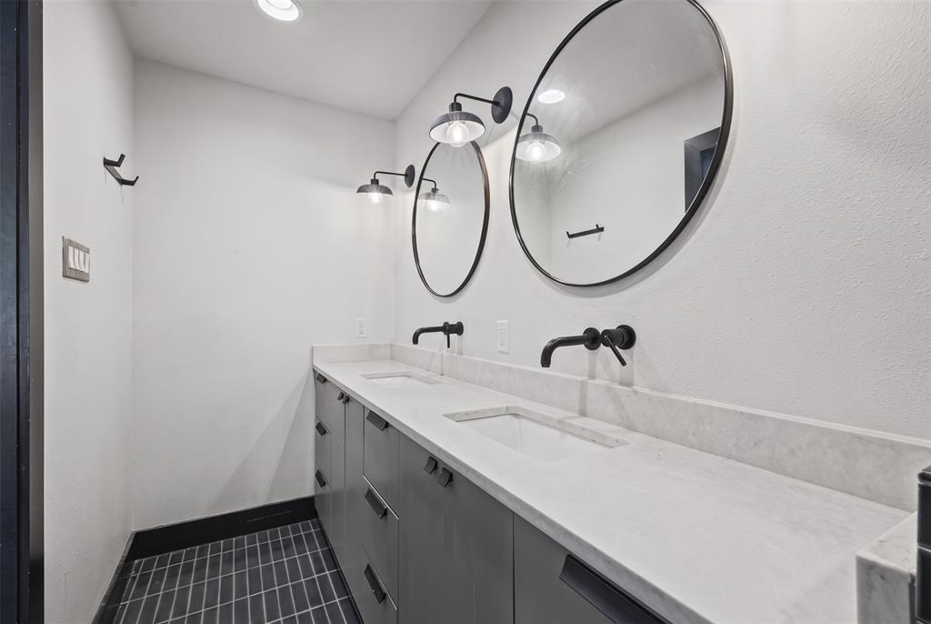 Dual-sink vanity featuring charcoal-finished cabinetry, a white stone countertop, wall-mounted matte black faucets, round mirrors, and industrial-style wall sconces