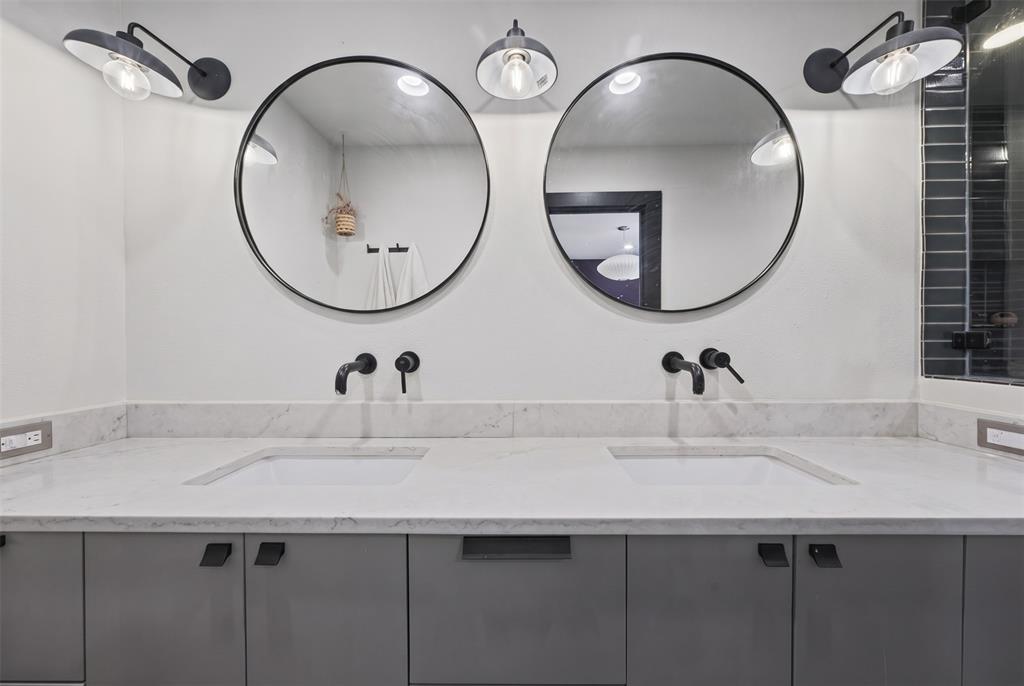 Dual-sink vanity featuring gray flat-panel cabinetry, light-toned stone countertops with integrated rectangular basins, matte black wall-mounted faucets, dual round mirrors, and matching industrial-style sconces