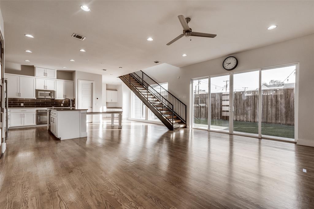 Open-concept interior featuring wood-finish flooring, recessed lighting, a kitchen with white cabinetry and tiled backsplash, a staircase with metal railings, and sliding glass doors