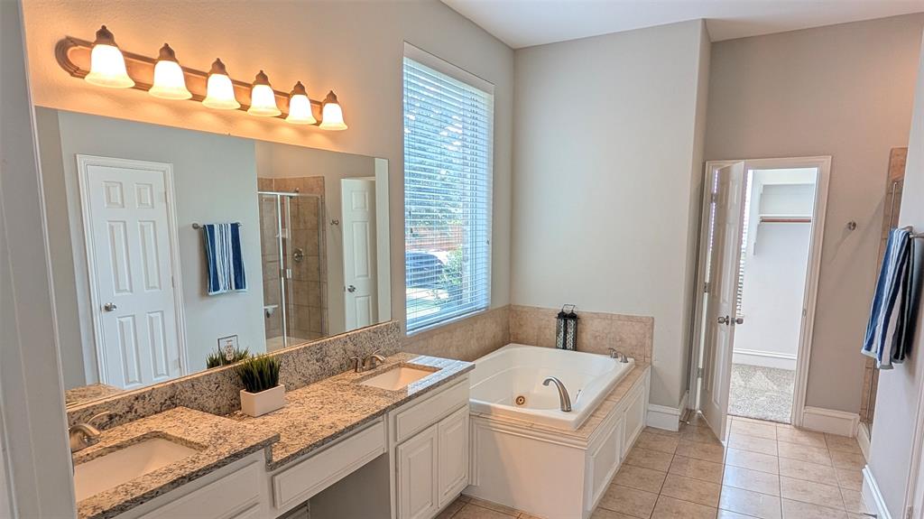 Dual-sink vanity with stone-finish countertops, soaking tub with tile surround, large wall-mounted mirror, and tile flooring