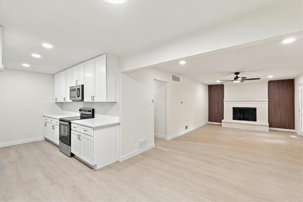 Kitchen featuring white shaker-style cabinetry, solid-surface countertops, and stainless steel appliances, accented by wood-finish flooring and a white painted brick fireplace