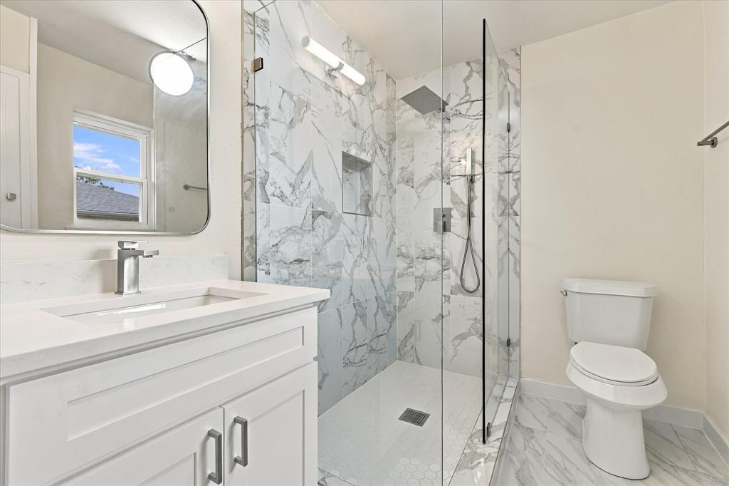 Bathroom featuring a stone-patterned tile walk-in shower with a built-in recessed niche, rainfall showerhead, glass enclosure, and white vanity with a stone countertop