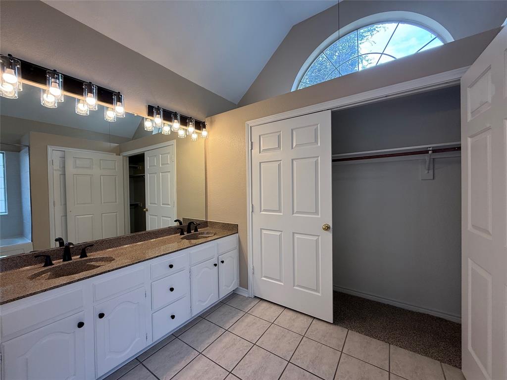 Double vanity featuring a stone-look countertop, dual undermount sinks, white cabinetry, tile flooring, and a vaulted ceiling with an arched window