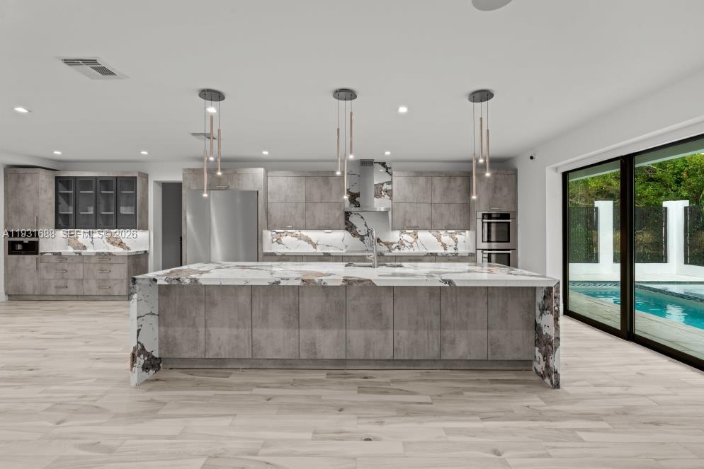 Kitchen featuring custom grey cabinetry, a waterfall-edge stone island with an integrated sink, stone slab backsplash, stainless steel built-in appliances, and wood-finish flooring