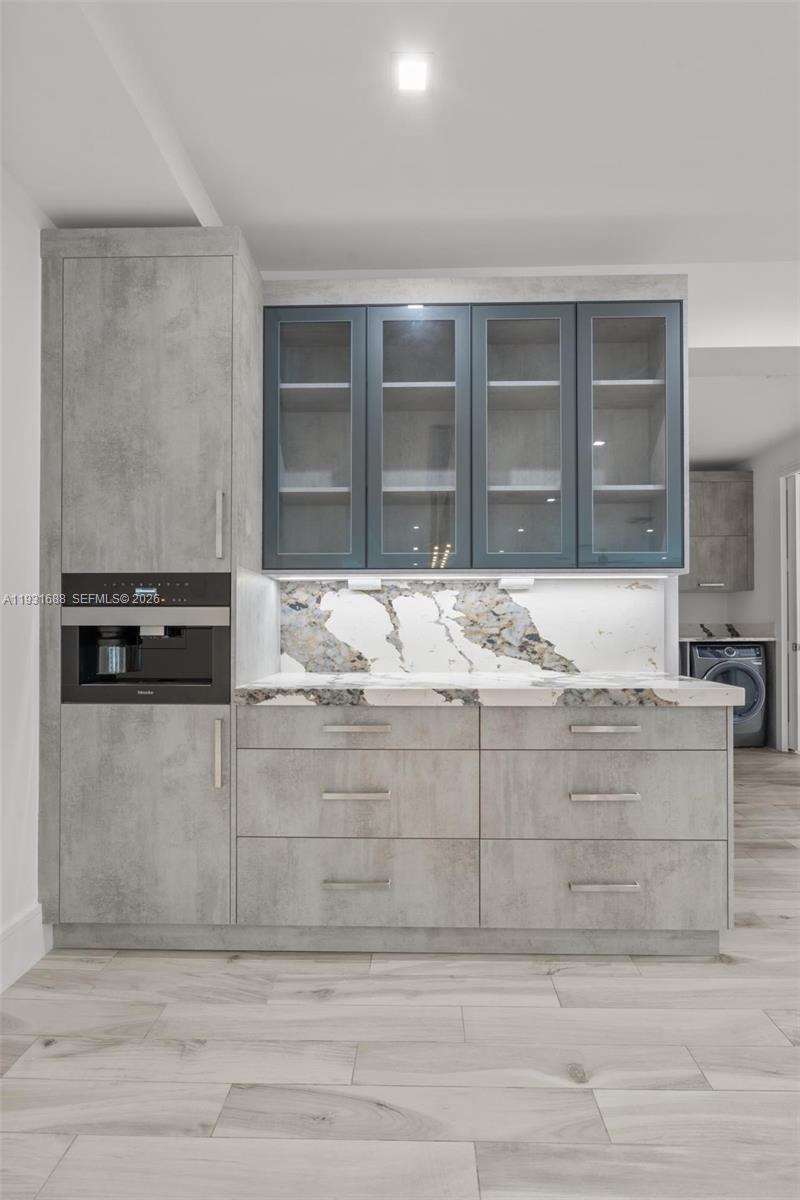 Custom cabinetry featuring grey textured finish and glass-front upper storage, stone-patterned backsplash, quartz countertop, and integrated built-in coffee station