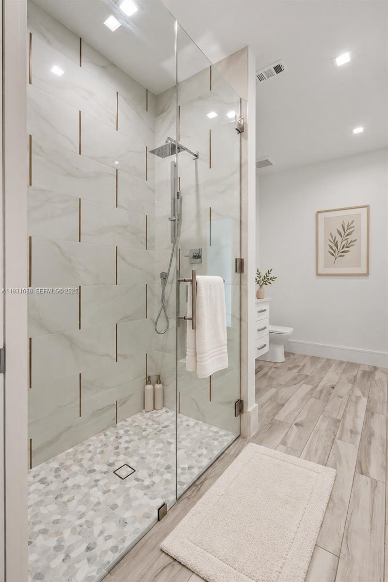 Walk-in shower featuring large-format tile walls with metallic inlay, river stone tile flooring, frameless glass enclosure, rainfall shower head, and wood-finish flooring