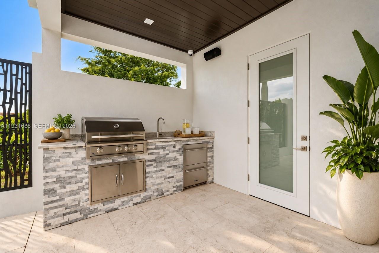 Covered outdoor kitchen featuring a stacked stone veneer base, built-in stainless steel grill, integrated storage, and a sink with a metal faucet, all situated on neutral-toned paved flooring