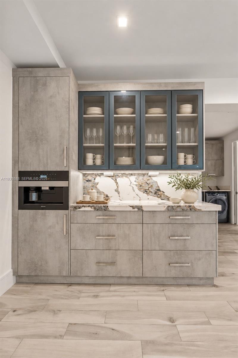 Textured grey cabinetry featuring glass-front upper displays, stone-patterned backsplash and countertop, integrated coffee system, and wood-finish tile flooring
