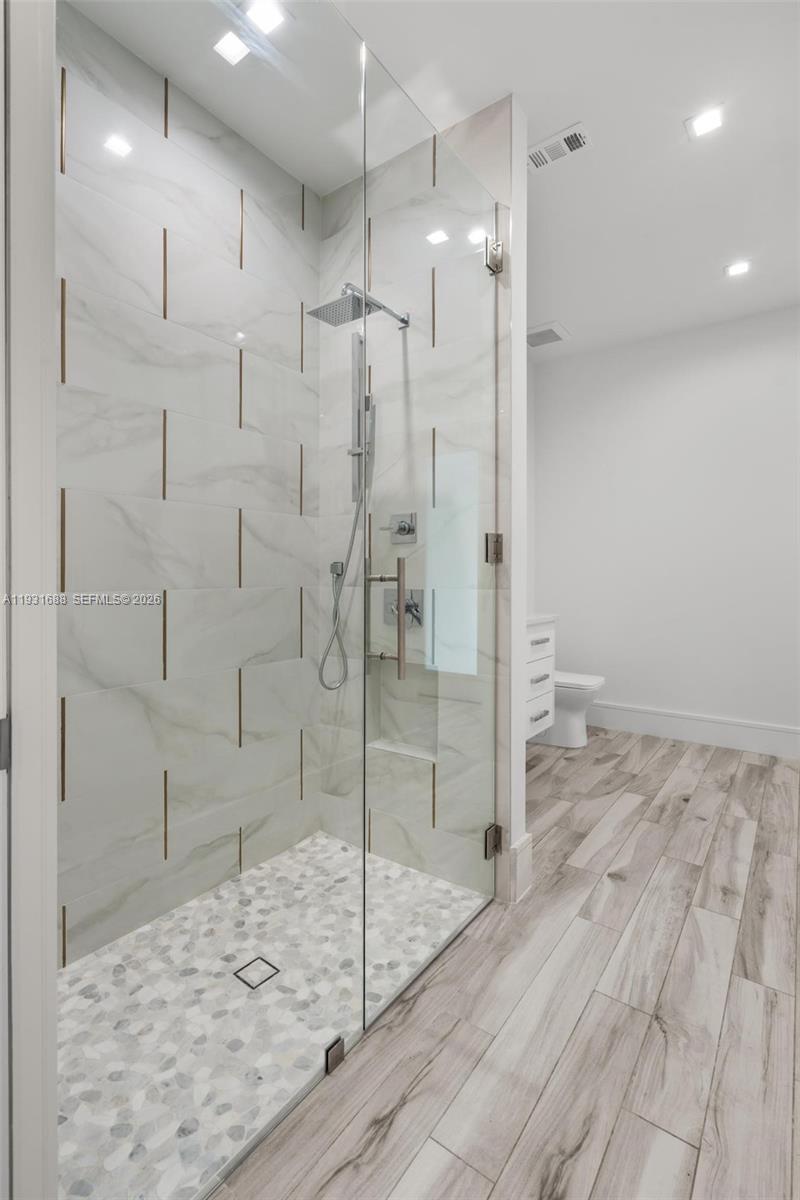Walk-in shower featuring large-format wall tile with metallic inlay, river stone mosaic floor, frameless glass enclosure, and rainfall showerhead, complemented by wood-finish flooring