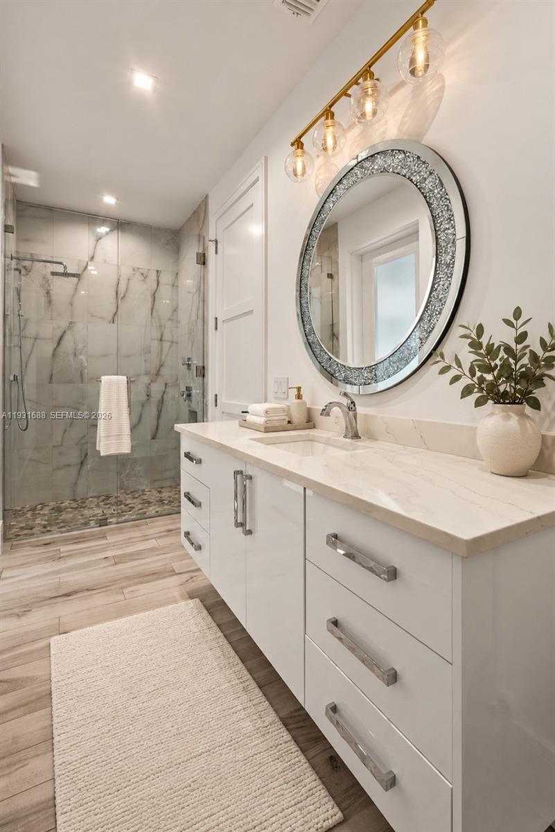 Bathroom featuring a white gloss vanity with stone-finish countertop, walk-in shower with stone tile surround and pebble floor, wood-finish flooring, and wall-mounted multi-light fixture