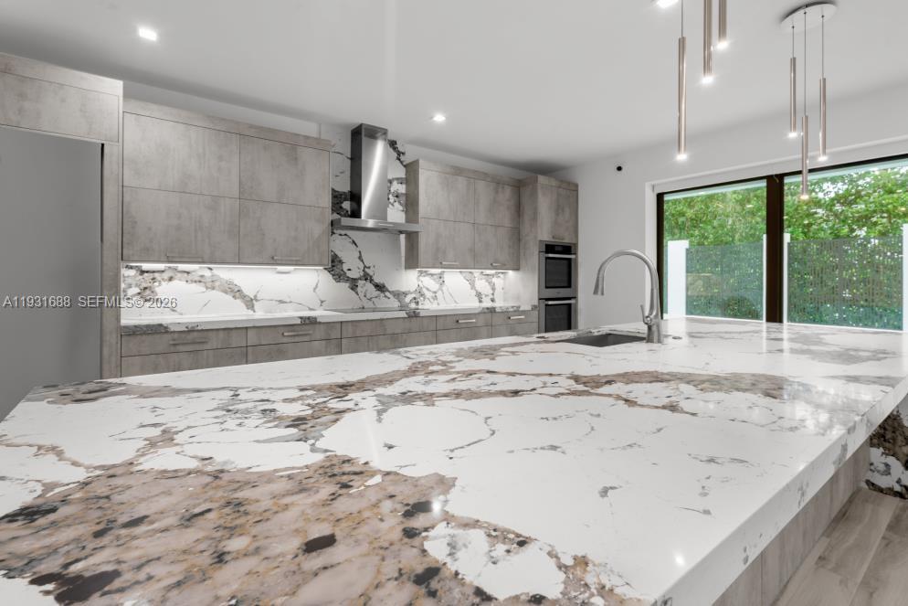 Kitchen featuring stone-patterned countertops and full-height backsplash, matte-finish cabinetry, built-in double wall ovens, stainless steel range hood, and pendant lighting