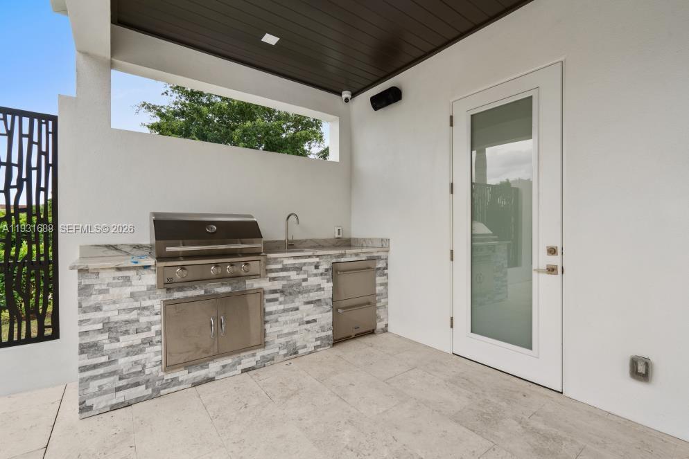 Covered outdoor kitchen featuring stone-veneer cabinetry, stone countertops, a stainless steel built-in grill, an under-counter refrigerator, and a stainless steel sink