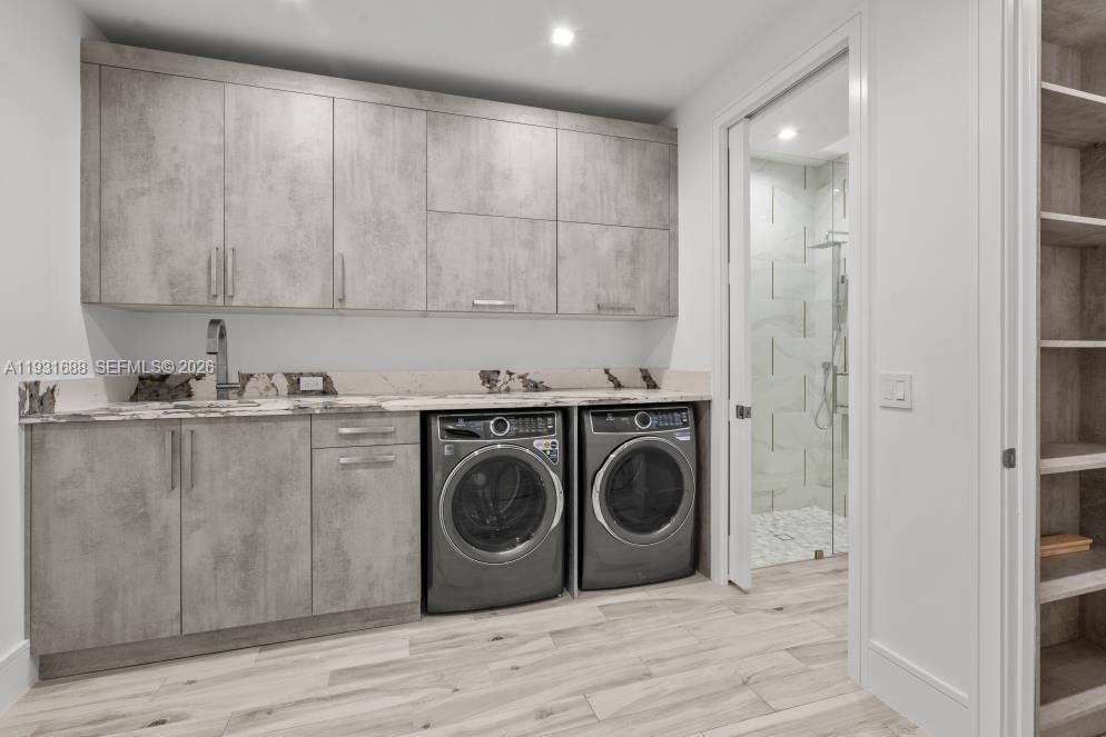 Laundry room featuring grey-toned cabinetry, stone-surface countertop with integrated sink, wood-finish flooring, and built-in shelving, with a glass-enclosed shower stall visible through the doorway