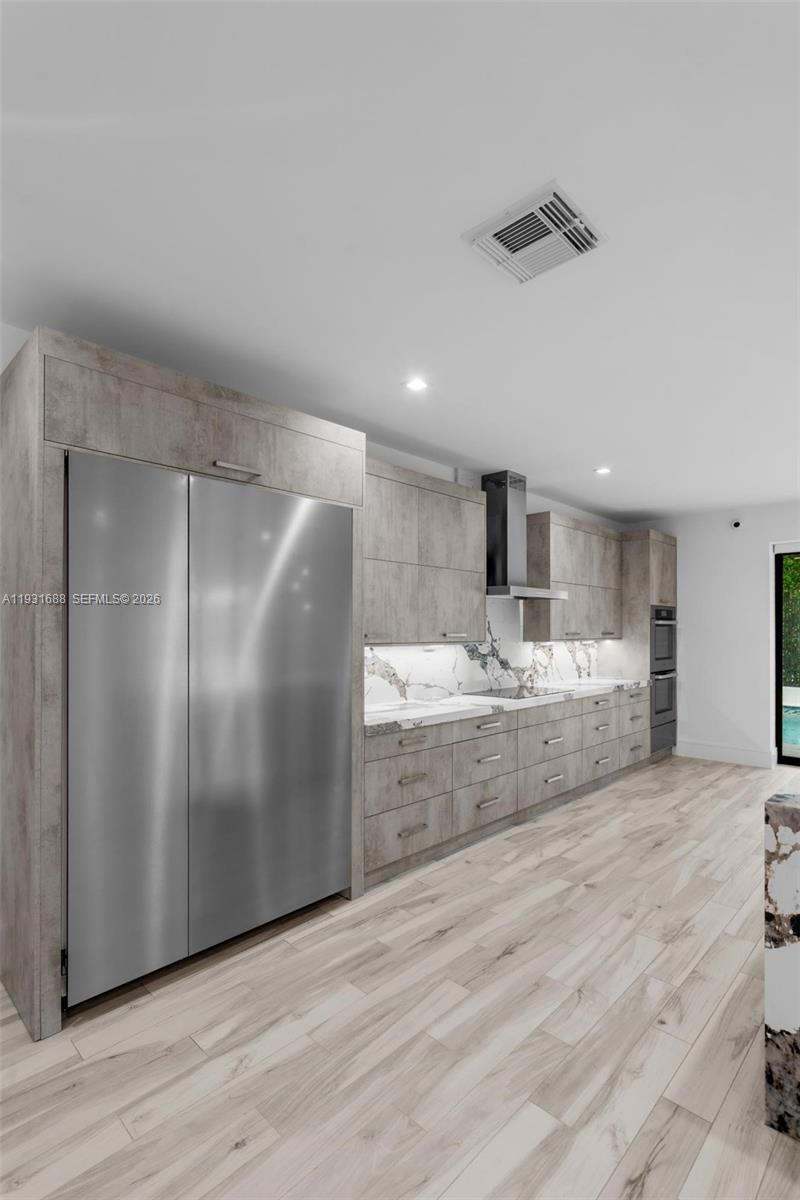 Kitchen featuring wood-finish cabinetry, integrated stainless steel refrigerator, stone-patterned backsplash, built-in double oven, and wood-finish flooring