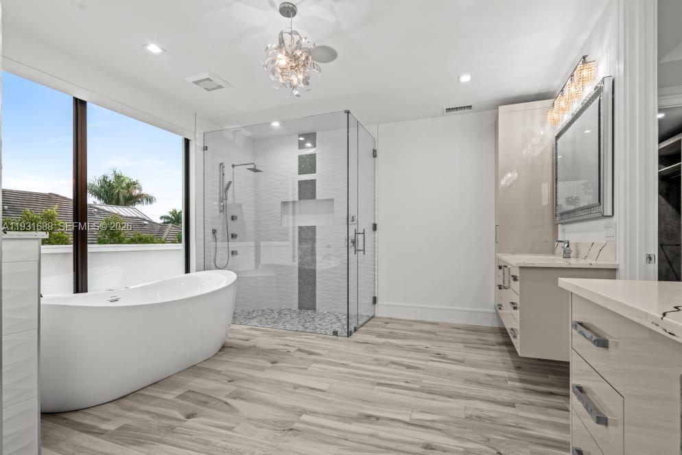 Freestanding soaking tub, glass-enclosed shower with tiled accents, custom cabinetry with stone countertops, and wood-finish flooring