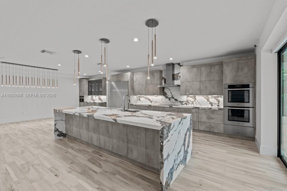 Kitchen featuring flat-panel cabinetry, a waterfall-edge island, stone-patterned counters and backsplash, stainless steel double wall ovens, and recessed lighting
