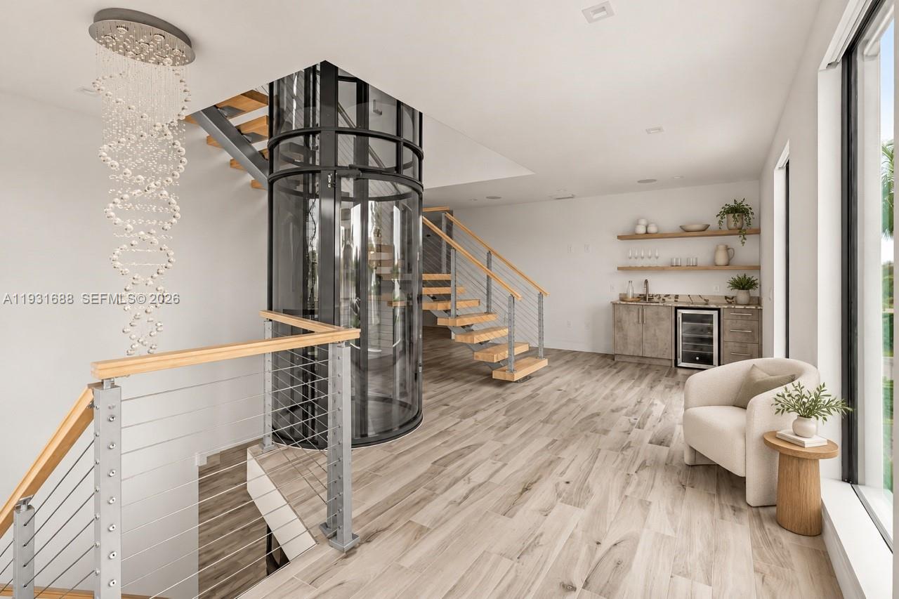 Cylindrical glass elevator, open-riser staircase with metal cable railing, wood-finish flooring, built-in wet bar with beverage cooler, and tiered wall shelving