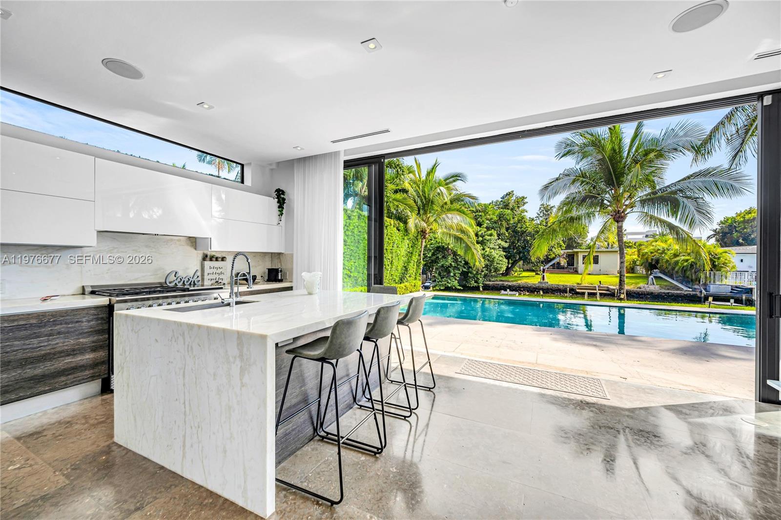 Modern kitchen featuring a large stone-topped waterfall island, high-gloss cabinetry, recessed lighting, and retractable floor-to-ceiling glass wall panels opening to a swimming pool and patio