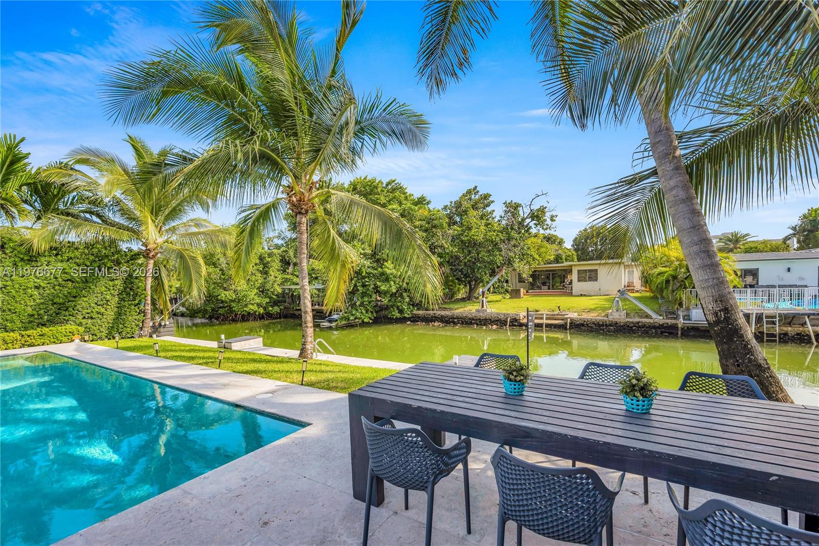 Waterfront backyard featuring a swimming pool, concrete patio, seawall, and mature palm trees