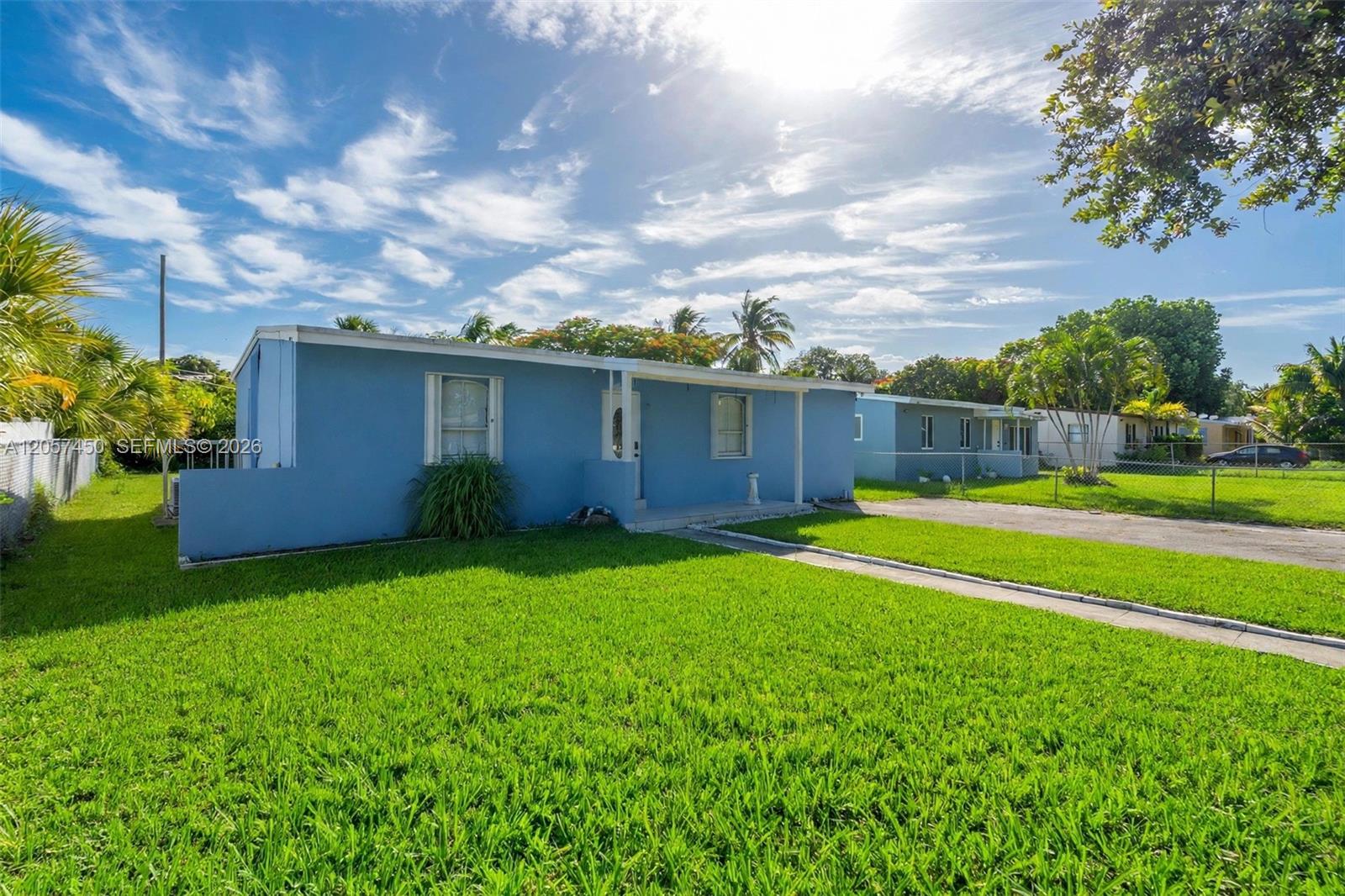 Single-story exterior featuring a blue facade, expansive front lawn, paved driveway, and mature palm trees