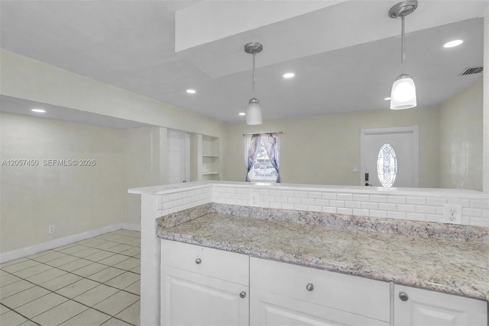 Kitchen featuring a stone-look countertop, white subway tile backsplash, white shaker-style cabinetry, two brushed-nickel pendant lights, and tile flooring
