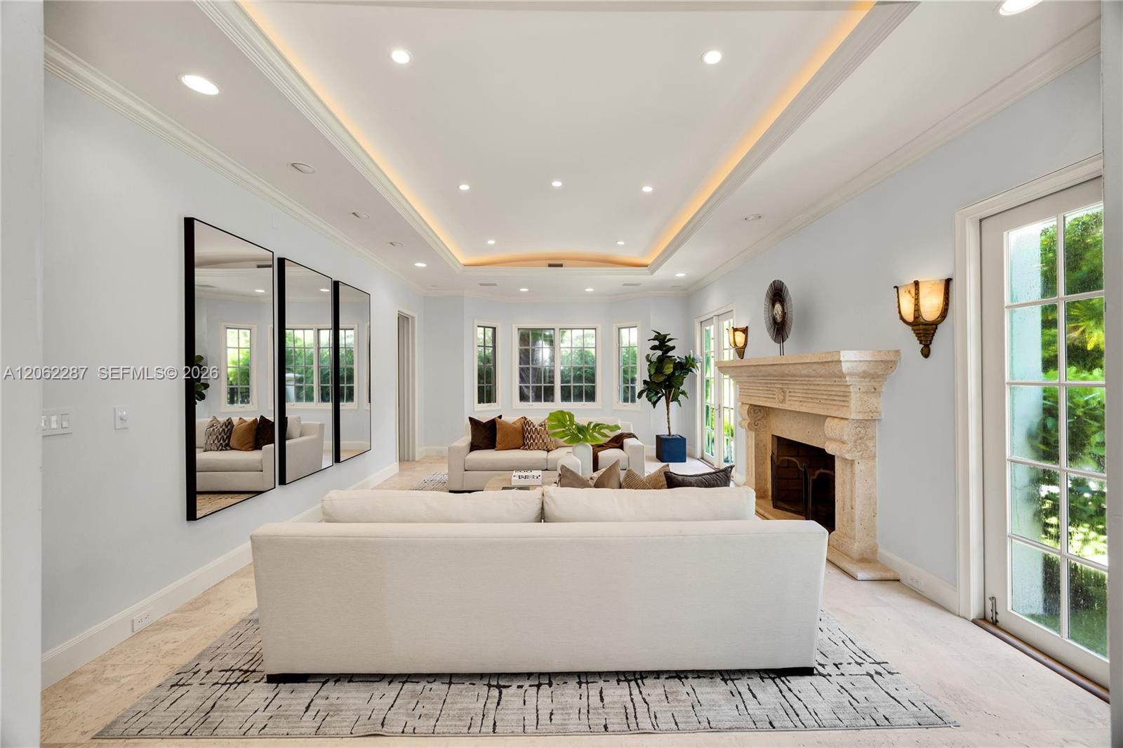 Living area featuring a tray ceiling with recessed lighting, carved stone fireplace mantel, wall-to-wall mirrors, light-toned stone flooring, and multiple sets of glass-paned doors