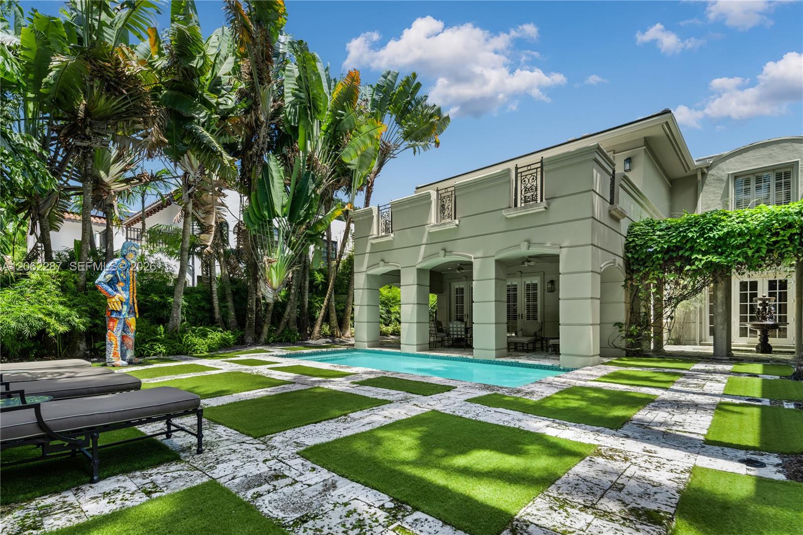 Rear exterior featuring a covered patio, rectangular swimming pool, stone-paver hardscaping with inset turf, and mature tropical landscaping