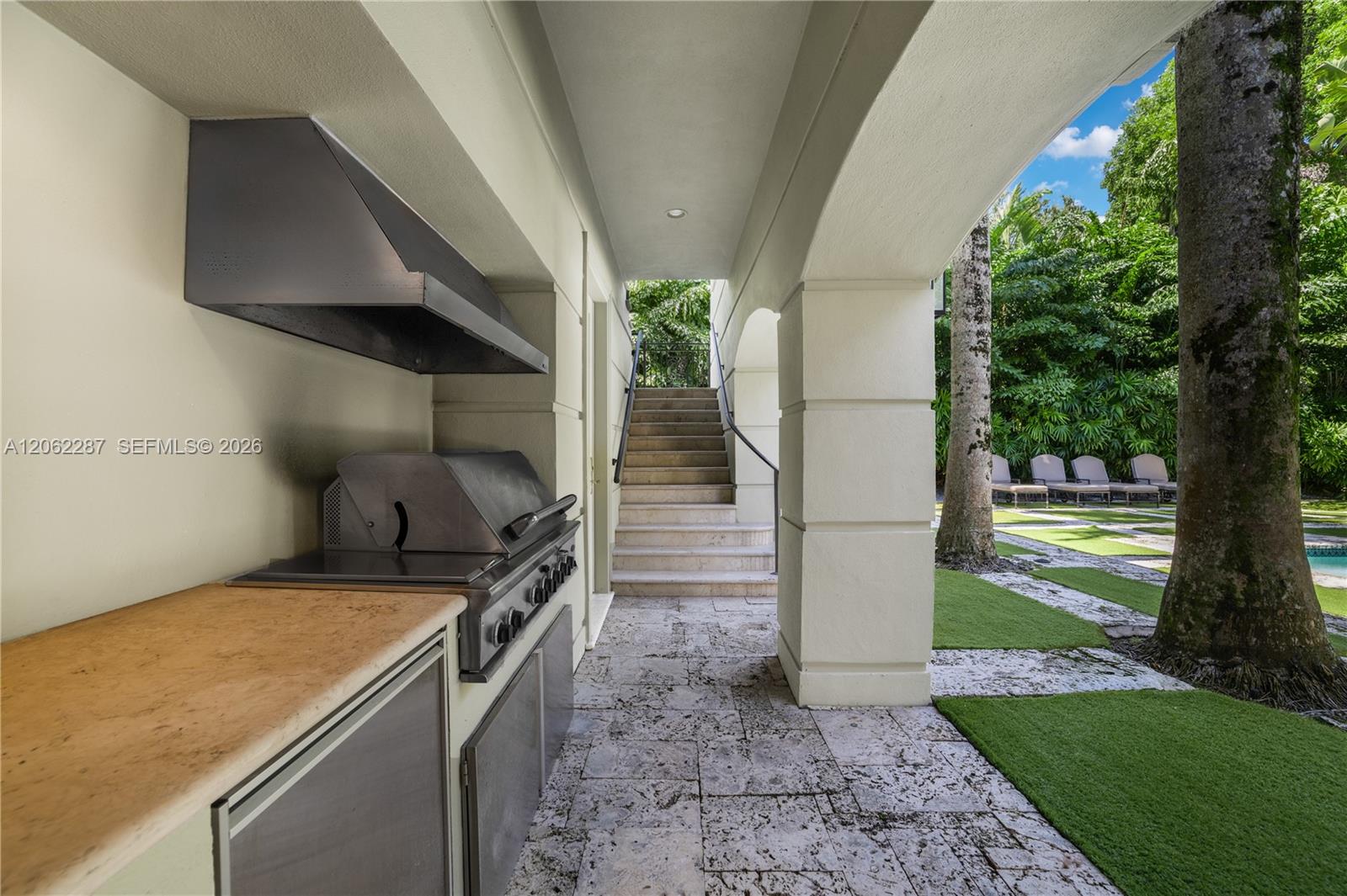 Covered outdoor kitchen featuring a built-in stainless steel grill, range hood, and solid surface countertop, complemented by stone tile flooring, arched architectural columns, and stone stairways