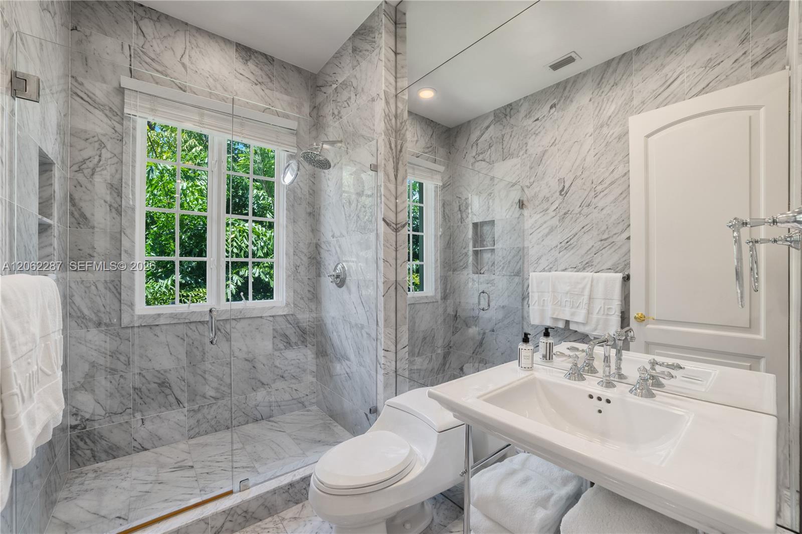Full-wall natural stone tiling, glass-enclosed shower, wall-to-wall vanity mirror, porcelain sink with polished chrome widespread faucet, and multi-pane window