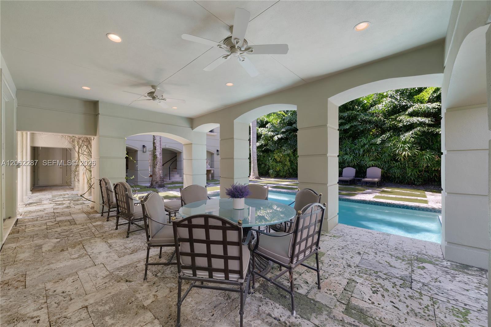 Covered outdoor loggia featuring stone paver flooring, architectural columns with arched openings, recessed lighting, and ceiling fans, overlooking a pool