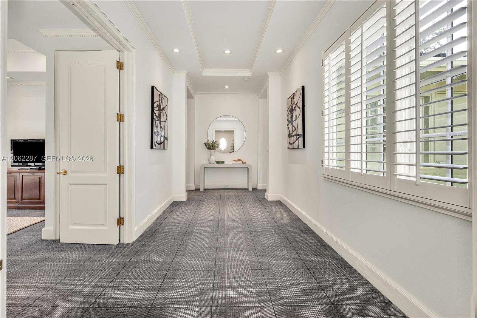 Hallway featuring recessed ceiling lighting, tray ceiling details, plantation shutters, and patterned carpeting