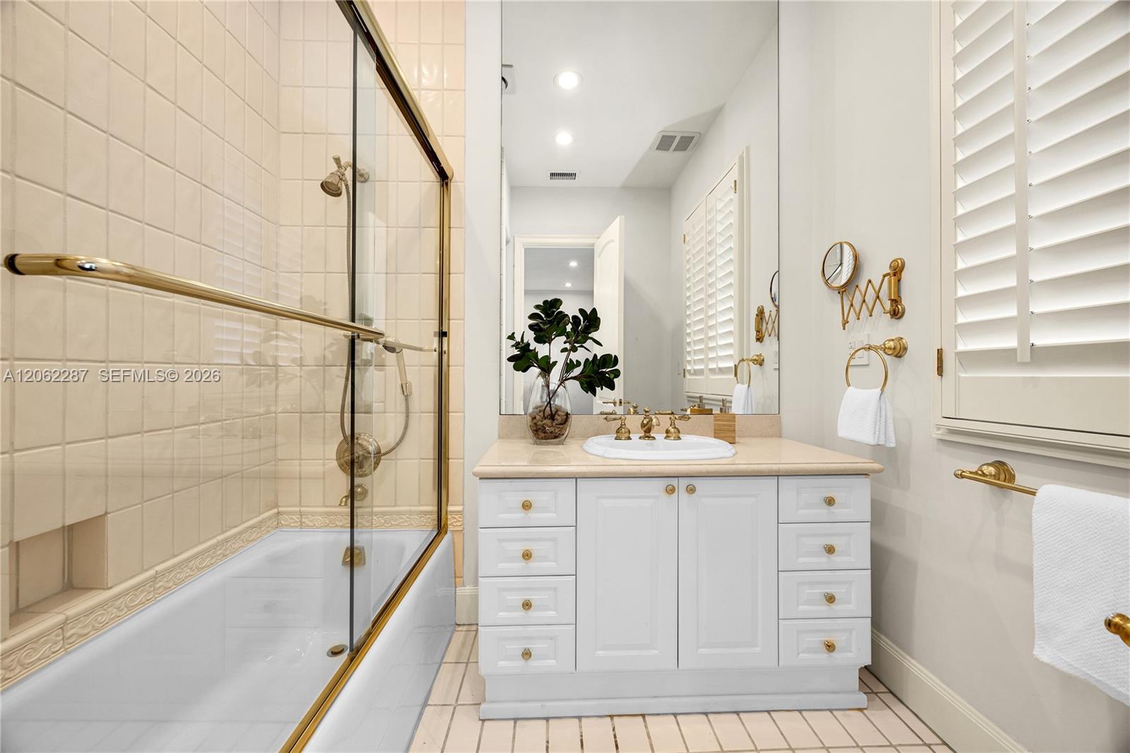 White vanity with stone countertop, undermount sink, brass-finish fixtures, and a glass-enclosed shower with tiled surround and plantation shutters