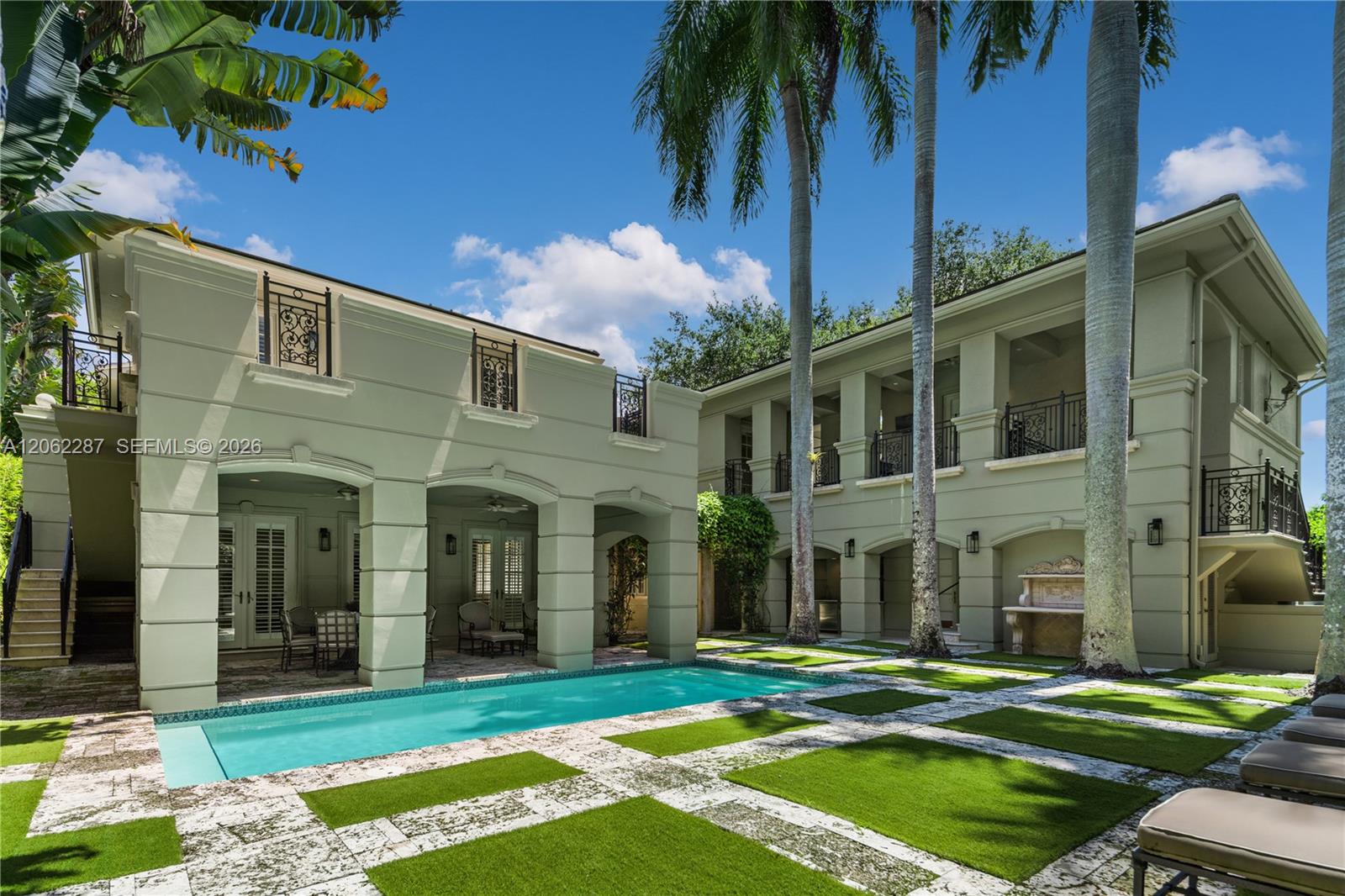 Two-story residence featuring a rectangular swimming pool, integrated covered loggias, ornamental iron balcony railings, stone-paver decking with turf accents, and mature palm trees