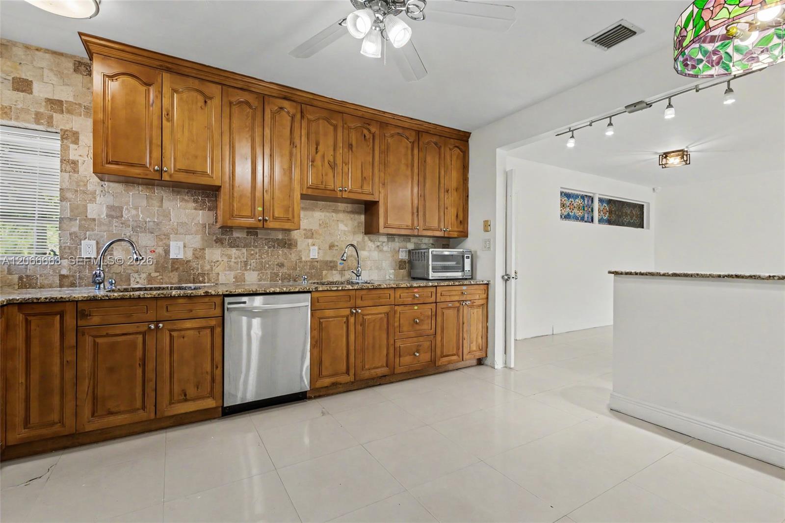 Kitchen featuring wood cabinetry, natural stone backsplash, stone countertops, a stainless steel dishwasher, and tile flooring