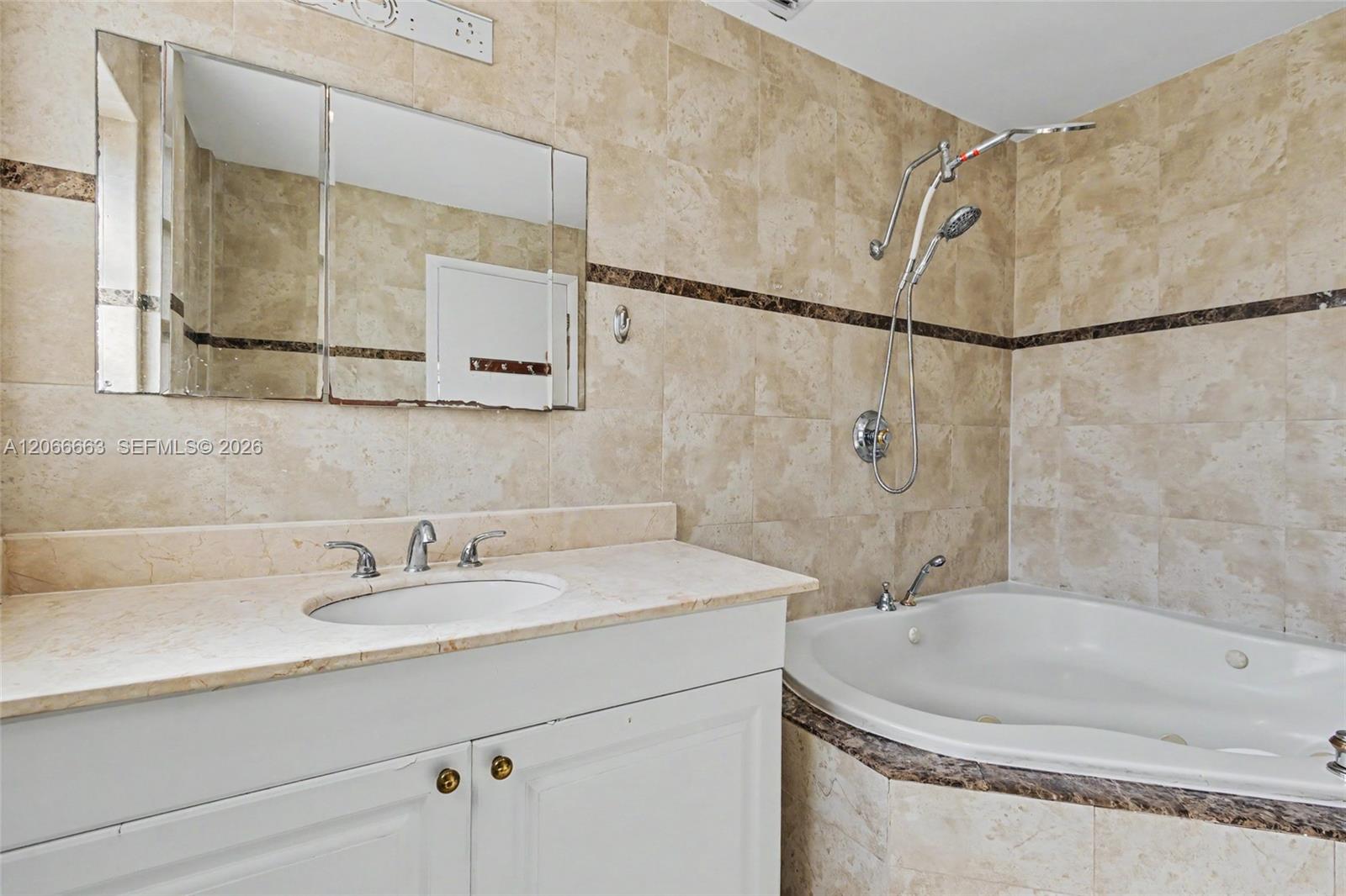 Bathroom featuring a white vanity cabinet, light stone countertop with an integrated basin, triple-panel mirrored medicine cabinet, built-in jetted tub, and wall tiling with decorative border accent