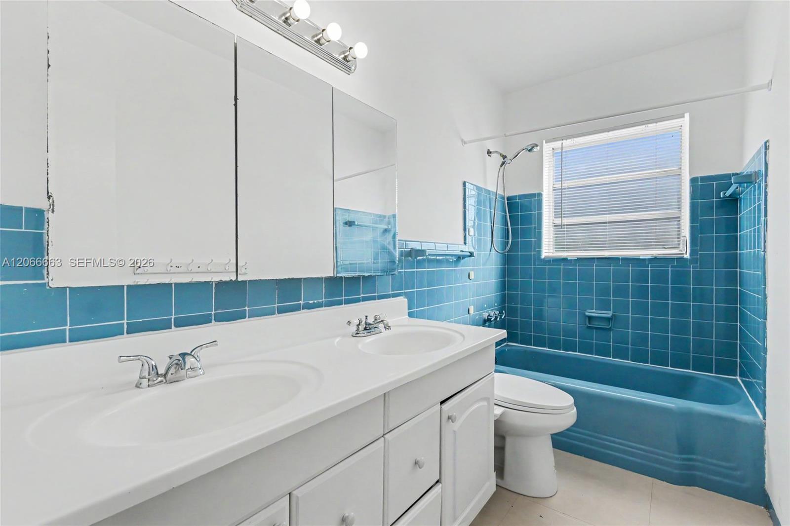 Dual-sink vanity with white countertop, integrated medicine cabinet mirrors, multi-bulb wall lighting, and blue tile wainscoting surrounding the matching blue bathtub