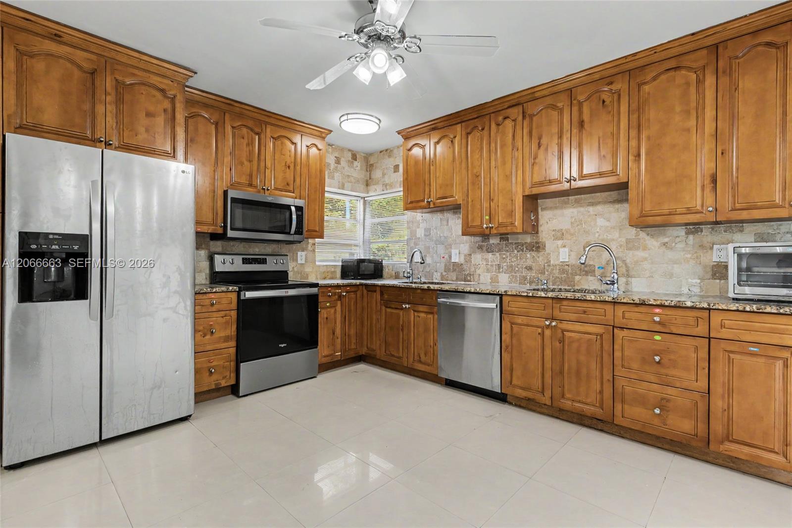 Kitchen featuring extensive wood cabinetry, stone-tiled backsplash, polished stone countertops, stainless steel appliances, and large-format tile flooring