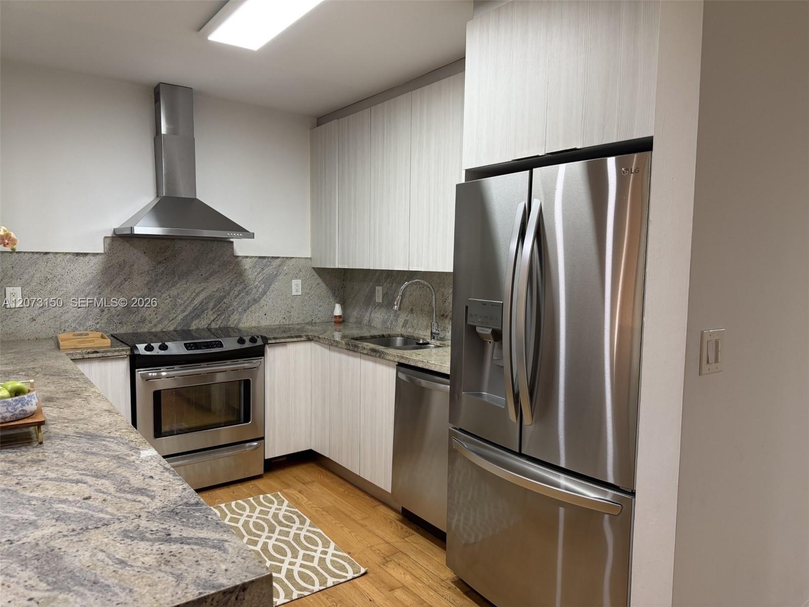 Kitchen featuring flat-panel cabinetry, stone countertops, stainless steel appliances, a mounted range hood, and wood-finish flooring
